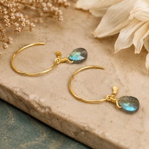 May include: A pair of gold hoop earrings featuring teardrop-shaped labradorite gemstones. Each earring has a thin gold wire hoop with a small, faceted, blue-grey labradorite stone attached by a wrapped gold wire detail at the bottom.