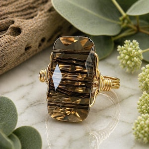 May include: A rectangular faceted smoky quartz gemstone ring featuring a gold-toned wire-wrapped band. The stone displays deep brown hues with visible dark needle-like inclusions throughout the translucent crystal structure of the ring.