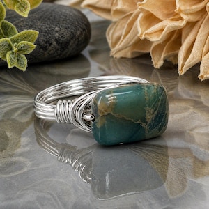 May include: A silver wire-wrapped ring featuring a rectangular, teal-colored stone with brown and gold marbling. The ring's band is composed of multiple strands of silver wire, creating a textured appearance. The stone is the focal point, set prominently on the ring.