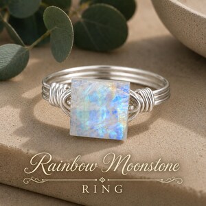 May include: A silver wire-wrapped ring featuring a square-cut rainbow moonstone with iridescent blue and white flashes. The band is constructed from multiple strands of silver wire coiled at the sides to secure the central gemstone setting.