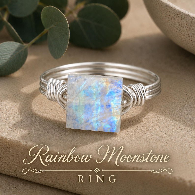 May include: A silver wire-wrapped ring featuring a square-cut rainbow moonstone with iridescent blue and white flashes. The band is constructed from multiple strands of silver wire coiled at the sides to secure the central gemstone setting.