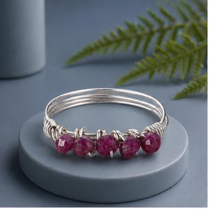 May include: A silver wire-wrapped bangle bracelet featuring five faceted, deep pinkish-red gemstone beads. The silver wire is coiled around the band to secure the stones in a horizontal row, creating a multi-layered metallic structure.