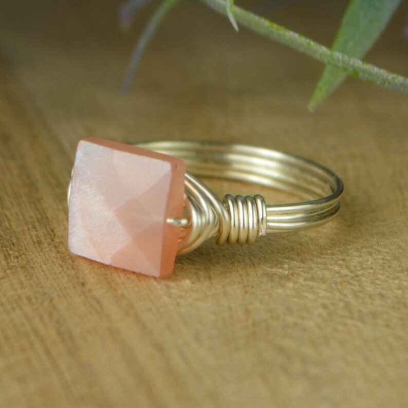May include: A silver wire-wrapped ring with a square, faceted, peach-colored gemstone. The ring has a multi-strand band and a decorative wire wrap around the gemstone setting. The ring is displayed on a wooden surface.