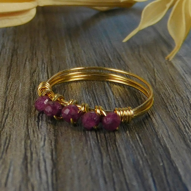 May include: A gold wire ring featuring a row of four small, faceted, deep pinkish-red gemstone beads. The band is constructed from multiple thin strands of gold-toned wire that are wrapped around the base of each bead to secure them in place.