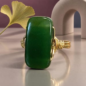May include: A ring featuring a large, rectangular, smooth dark green stone set in a gold-colored wire-wrapped band. The wire is coiled around the sides of the stone to secure it, creating a metallic frame that contrasts with the deep green surface.