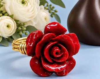 May include: A napkin ring featuring a glossy, deep red rose bloom attached to a gold-toned metal wire band. The rose has layered, curved petals with a smooth, reflective finish, while the band consists of multiple coiled strands of gold wire.