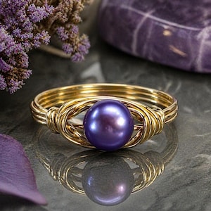 May include: A gold-toned wire ring featuring a large, round, iridescent purple pearl. The ring's design incorporates woven wire detailing around the pearl, creating a unique and elegant look. The ring is displayed on a reflective surface, showcasing its intricate design.