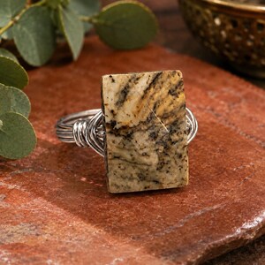 May include: A ring featuring a rectangular, tan and brown patterned stone set in a silver-toned wire-wrapped band. The stone displays natural dark veining and a smooth, flat surface, while the metal wire is coiled around the base of the setting.