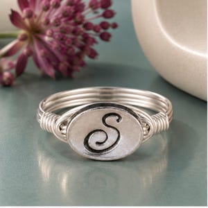 May include: A silver-toned ring featuring a central oval-shaped charm embossed with the letter S. The band is constructed from multiple thin, coiled silver wires that wrap around the sides of the oval centerpiece, creating a layered, textured appearance.
