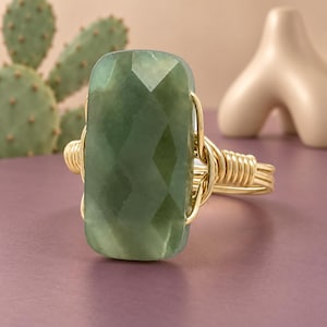 May include: A ring featuring a large, rectangular, faceted green gemstone set in a gold-colored wire-wrapped band. The stone has a smooth, polished surface with geometric facets that catch the light, secured by coiled gold wire on both sides.