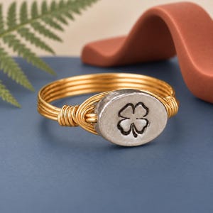 May include: A ring featuring a gold-toned wire band wrapped around an oval-shaped silver-toned metal centerpiece. The centerpiece is embossed with a four-leaf clover design. The band is constructed from multiple strands of thin gold-colored wire.