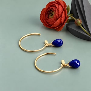 May include: A pair of gold hoop earrings featuring a single faceted, teardrop-shaped blue lapis lazuli gemstone dangling from each wire. The stones are secured with gold wire wrapping, and the hoops have a small flat tab at the end.