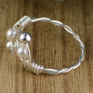 May include: A silver ring with a twisted band and a cluster of small, round, white pearls and silver beads. The ring is set against a wooden background, with a close-up view highlighting the details of the jewelry.