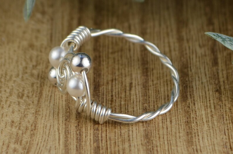 May include: A silver ring with a twisted band and a cluster of small, round, white pearls and silver beads. The ring is set against a wooden background, with a close-up view highlighting the details of the jewelry.