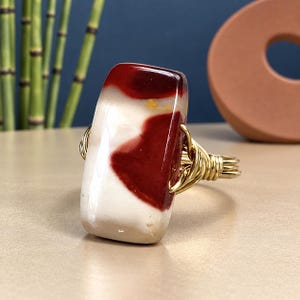 May include: A ring featuring a rectangular, polished stone with a smooth surface patterned in cream and deep red. The stone is secured by a gold-colored wire-wrapped band that coils around the sides of the stone to hold it firmly in place.