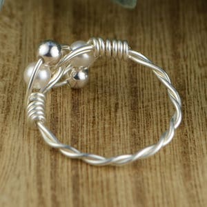 May include: A silver-toned ring with a twisted band and a cluster of small, round beads. The beads include both shiny silver and white pearl-like elements. The ring is set against a wooden surface.