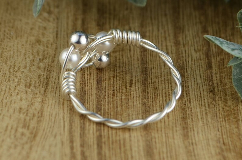 May include: A silver-toned ring with a twisted band and a cluster of small, round beads. The beads include both shiny silver and white pearl-like elements. The ring is set against a wooden surface.