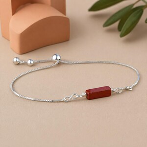 May include: A silver chain bracelet featuring a rectangular, deep red stone bead at the center. The bracelet includes a sliding mechanism with two small silver spheres at the ends of the chain, allowing for adjustable sizing on the wrist.