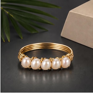 Pode incluir: Uma pulseira rígida de fio dourado com cinco pérolas brancas lisas de formato oval dispostas em fila na parte frontal. A pulseira é composta por vários fios metálicos finos e enrolados que envolvem as pérolas.