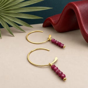 May include: A pair of gold hoop earrings featuring a dangling charm composed of four small, faceted, deep pink ruby-colored beads. The beads are arranged in a vertical line, finished with a small gold bead at the bottom of each earring.