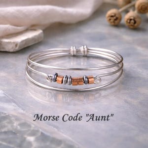 May include: A silver wire bracelet with three bands, featuring copper and silver beads spelling "Aunt" in Morse code. The bracelet is displayed on a reflective surface, with the text "Morse Code 'Aunt'" below.