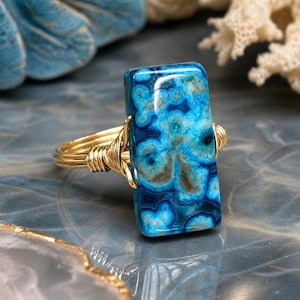 May include: A gold-toned wire-wrapped ring featuring a rectangular blue gemstone with swirling patterns of teal and brown. The ring's band is a simple, polished gold. The gemstone is approximately 1 inch long.