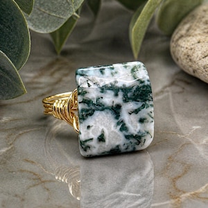 May include: A gold-colored wire-wrapped ring featuring a square-cut gemstone. The gemstone is white with green and dark green veining. The ring is set against a marble-like surface with greenery in the background.