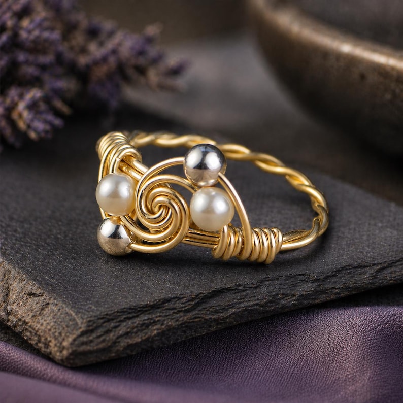 May include: A gold-colored wire-wrapped ring featuring a spiral design with two faux pearls and two silver-colored beads. The ring is set on a dark gray stone surface, with a blurred purple background.