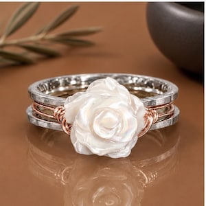 May include: A ring featuring a central white carved floral element, likely mother-of-pearl, secured by copper-coloured wire wrapping. The band consists of two outer silver-toned hammered metal rings and a central copper-coloured wire band.