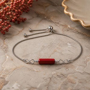May include: A silver-toned bracelet with a red rectangular center stone. The bracelet features an adjustable chain and infinity symbol accents on either side of the stone. The background includes a stone surface and decorative elements.