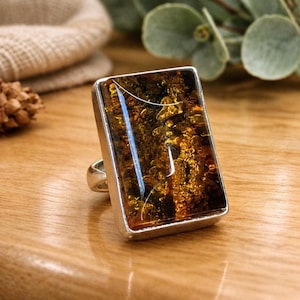 May include: A rectangular ring with a large, amber-colored stone set in a silver-toned metal frame. The stone has a rich, golden-brown hue with intricate patterns. The ring is displayed on a wooden surface.