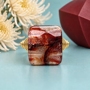 May include: A ring featuring a rectangular, polished stone with deep red, brown, and white marbled inclusions. The stone is set in a gold-toned wire-wrapped band that coils around the sides of the setting, creating a textured, metallic finish.