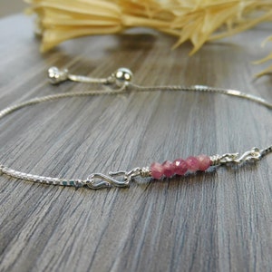 May include: A silver chain bracelet with a pink gemstone accent. The bracelet has a sliding clasp for adjustable sizing.