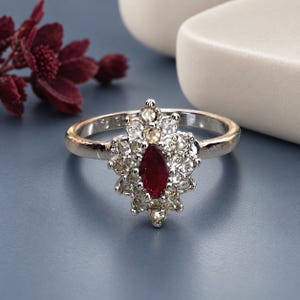 May include: A silver-toned ring featuring a central marquise-cut deep red gemstone surrounded by a cluster of small, clear, round-cut stones arranged in a pointed, elongated floral-inspired shape on a polished metal band.