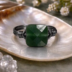 May include: A handcrafted ring featuring a faceted, square-cut green gemstone set in a dark wire wrap. The ring's band is made of multiple strands of dark wire. The ring is displayed on a white, iridescent surface.