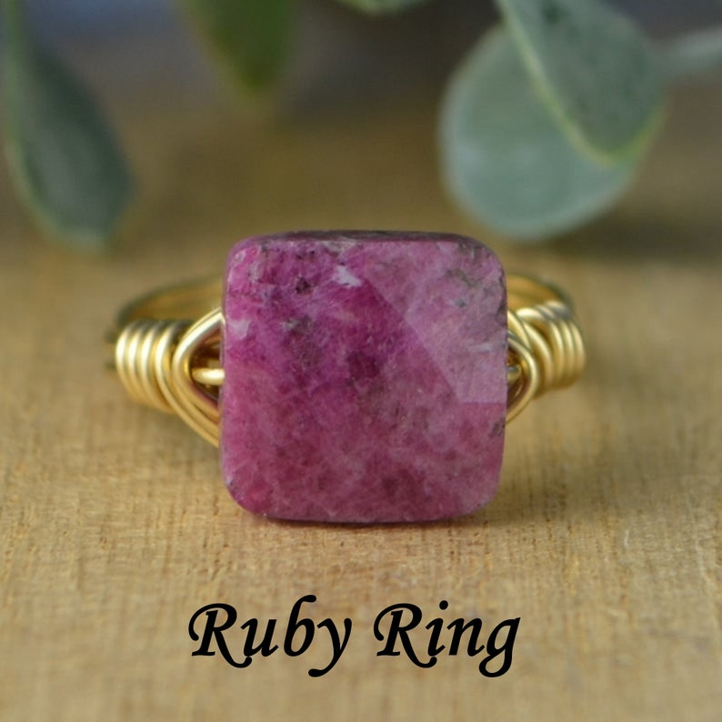 May include: A gold-toned wire-wrapped ring featuring a square, faceted, deep pink ruby gemstone. The ring is set against a blurred wooden background with the text "Ruby Ring" in a script font.