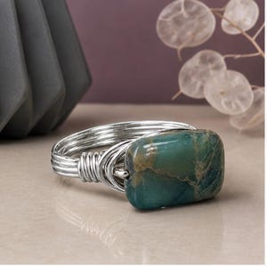 May include: A ring featuring a rectangular, teal-colored stone with brown veining, set into a band constructed from multiple strands of silver-toned wire that are wrapped and coiled together at the sides to secure the stone in place.
