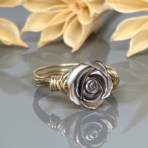 May include: A silver rose ring with a gold wire band. The rose is made of metal and has a detailed design.