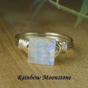 May include: A silver wire-wrapped ring featuring a square rainbow moonstone. The moonstone displays iridescent colors. The text "Rainbow Moonstone" is below the ring.