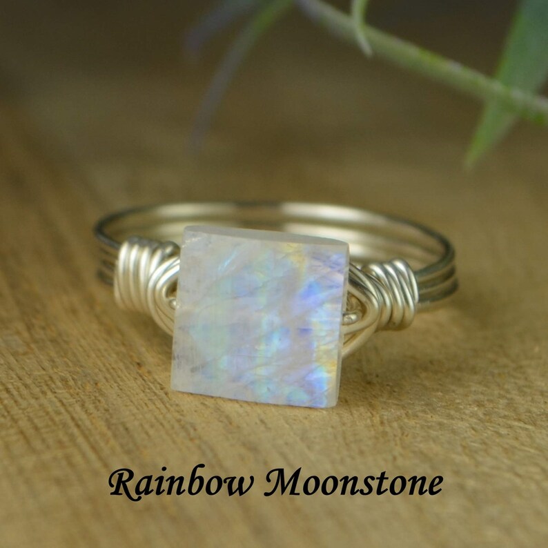 May include: A silver wire-wrapped ring featuring a square rainbow moonstone. The moonstone displays iridescent colors. The text "Rainbow Moonstone" is below the ring.