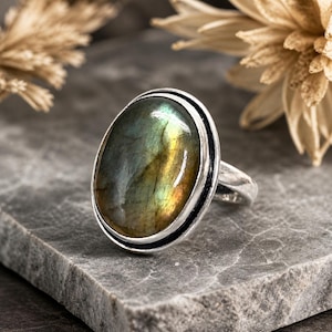 May include: A silver ring featuring an oval labradorite gemstone. The gemstone displays iridescent colors of green, yellow, and blue. The ring is set on a gray stone surface, with dried flowers in the background.