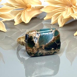 Peut inclure: Bague artisanale ornée d'une pierre rectangulaire de couleur teal avec des veines marron et dorées. La pierre est sertie dans un fil d'or. La bague est présentée sur une surface réfléchissante avec des fleurs séchées en arrière-plan.
