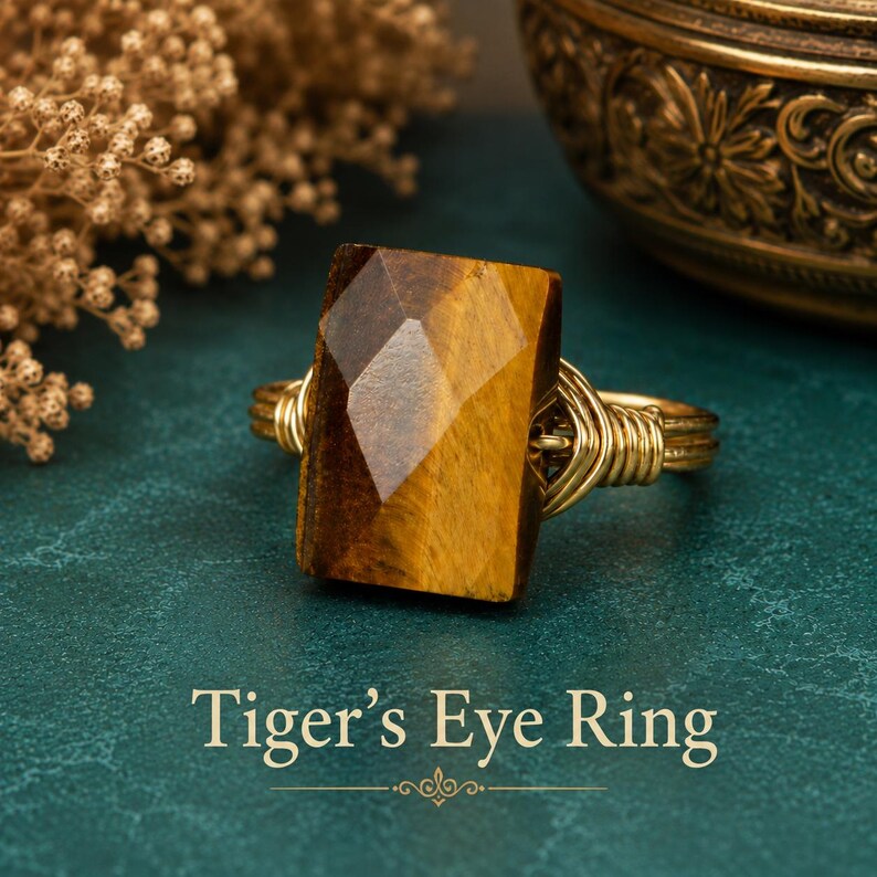 May include: A ring featuring a rectangular, faceted tiger's eye gemstone with brown and gold bands, secured by a coiled gold-toned wire band. The text "Tiger's Eye Ring" is displayed at the bottom of the image in a serif font.