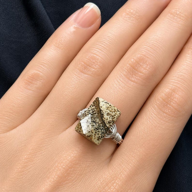 May include: A wire-wrapped ring featuring a faceted, rectangular-shaped stone with a speckled beige and brown pattern. The band is constructed from thin, coiled silver-toned wire that secures the stone in place with multiple wraps around the base.