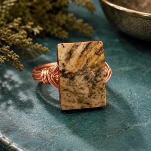 May include: A ring featuring a rectangular, tan-colored stone with dark brown and black dendritic inclusions. The stone is secured by a copper wire-wrapped band that coils around the base of the setting, creating a textured, metallic appearance.