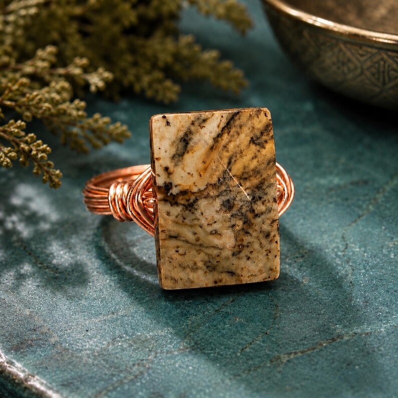 May include: A ring featuring a rectangular, tan-colored stone with dark brown and black dendritic inclusions. The stone is secured by a copper wire-wrapped band that coils around the base of the setting, creating a textured, metallic appearance.