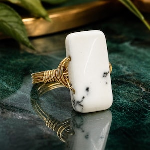 May include: A handcrafted ring featuring a rectangular white stone with black speckles, set in a gold-colored wire wrap. The ring is displayed on a reflective, dark green surface, showcasing its intricate design and craftsmanship.