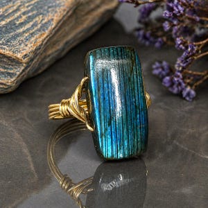 May include: A gold wire-wrapped ring featuring a rectangular, iridescent blue-green gemstone. The gemstone has vertical striations and is set against a dark background with a stone and purple flowers.