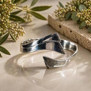May include: A silver-toned cuff bracelet with a geometric design. The bracelet has a curved band that wraps around the wrist, with two triangular accents that overlap. The bracelet is displayed on a neutral surface with greenery in the background.