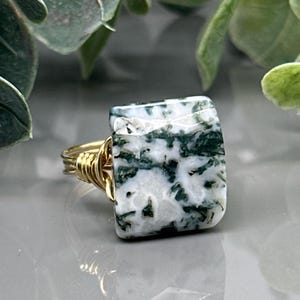 May include: A square-cut ring featuring a moss agate stone with white and green patterns. The stone is set in a gold-colored wire wrap band, creating a unique and naturalistic jewelry piece.